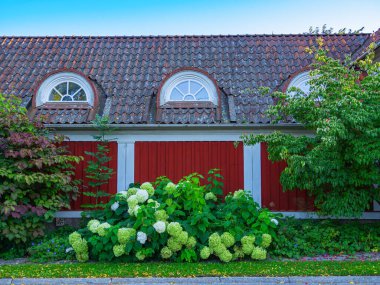 Stockholm, İsveç - 6 Eylül 2024: Yuvarlak pencereli ve yemyeşil bahçeli büyüleyici kırmızı ahşap ev.