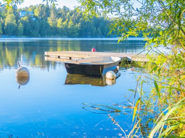 Bereketli yeşillik ve mavi gökyüzünü yansıtan durgun suyla çevrili ahşap rıhtıma bağlı küçük bir tekneyle sakin bir göl manzarası..