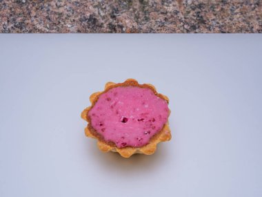 Pembe dolgulu tek bir tart, açık arkaplana yerleştirilmiş. Turtanın istiridyeli kenarı var ve yeni yapılmış gibi görünüyor. Dolgunun pürüzsüz bir dokusunu gösteriyor..