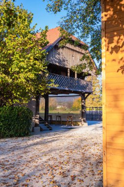 Domzale, Slovenya - 29 Ekim 2024: Balkonlu, yemyeşil ağaçlar ve yerdeki yapraklarla çevrili büyüleyici bir ahşap 2 katlı saman. Sahne parlak ve güneşli, sakin bir açık hava manzarası sergiliyor..