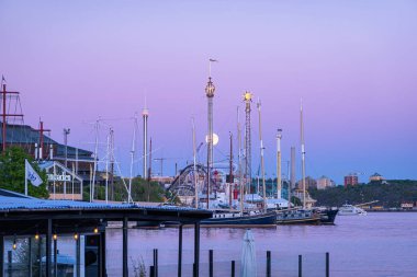 Stockholm, İsveç - 22 Mayıs 2024: Günbatımında sakin bir liman sahnesi, arka planda bir lunaparkta yükselen dolunay, sakin ve resimli bir atmosfer yaratıyor..