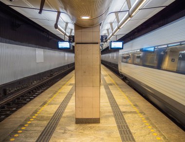 Kopenhag, Danimarka - 23 Mart 2025: Merkezi bir sütunun yer aldığı iki platformlu modern bir metro istasyonu. Bir tren hareket halinde, hız hissi yaratıyor. Dijital görüntüler tren bilgisini gösterir.