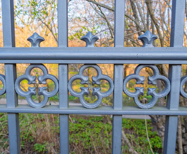 Dekoratif desenli süslü metal bir çitin yakın çekim görüntüsü. Çit karmaşık tasarımlar içeriyor ve bir ağaç ve yeşillik zeminine dayanıyor. Bu da sakin bir açık hava ortamı olduğunu gösteriyor..