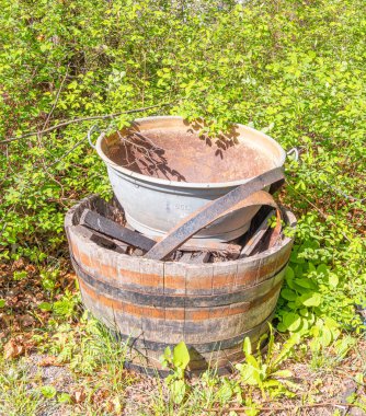 Ahşap bir fıçının üzerine yerleştirilmiş, yemyeşil yapraklarla çevrili eski bir metal kova. Sahne, doğal elementlerle birlikte kırsal bir açık hava görüntüsü yakalıyor..