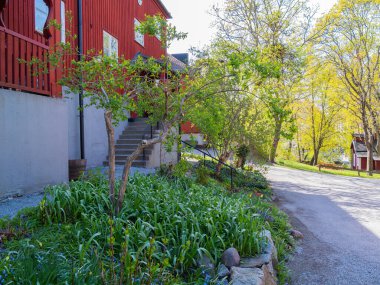 Yeşillik ve çiçek açan çiçeklerle çevrili merdiveni olan büyüleyici kırmızı bir evin manzarası. Güneş ışığı ağaçların arasından süzülüp sıcak ve davetkar bir atmosfer yaratır..