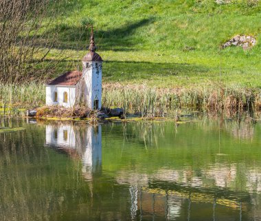 Bloke, Slovenya - 12 Nisan 2025: Çimen ve ağaçlarla çevrili sakin bir göl kenarındaki küçük, pitoresk bir şapel. Şapel kendine özgü bir çan kulesine sahiptir ve sakin sularda güzel bir şekilde yansıtılır..
