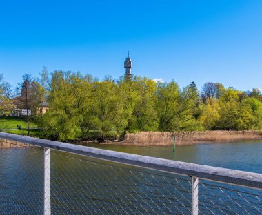 Sahilleri saran yemyeşil ağaçlarla dolu bir nehrin manzarası, arka planda bir kule ve açık mavi bir gökyüzü. Ön planda bir köprü parmaklığı var, perspektifi geliştiriyor..
