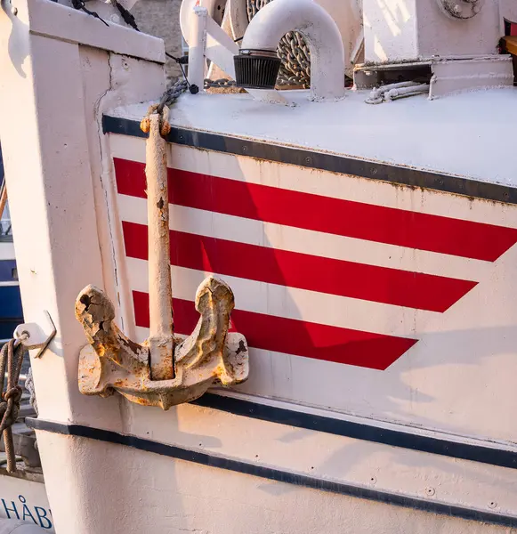 Eski beyaz bir çapa ve kırmızı beyaz çizgili bir dizayn içeren bir teknenin pruvasının yakın çekimi. Çapa, teknenin dokulu yüzeyine doğru belirgin bir şekilde sergileniyor. Bu da bir denizcilik temasını işaret ediyor..
