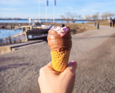 Kakaolu dondurma külahını tutan bir el, deniz manzaralı arka planda pembe kaplamalı..
