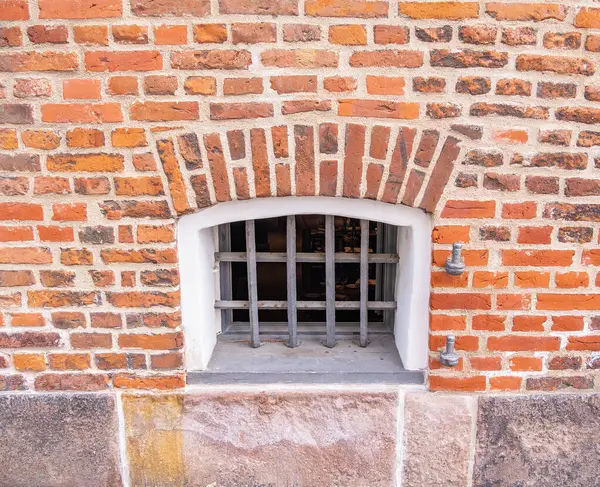 Küçük parmaklıklı pencereli tuğla bir duvar. Pencerenin metal bir ızgarası var ve taşralı bir temele kurulmuş. Tuğlalar kırmızı ve kahverengi renklerin karışımı, dokulu bir görünüm yaratıyor..