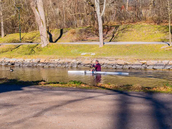 Stockholm, İsveç - 28 Nisan 2025 Dar bir su yolunda, ağaçlar ve çimenli bankalarla çevrili bir kürekçi. Sahne, güneş ışığının suya yansıdığı, huzurlu bir açık hava aktivitesi anı yakalıyor..