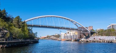 Stockholm, İsveç - 5 Mayıs 2025: Ağaçlar ve kentsel binalarla çevrili sakin bir nehir üzerinde uzanan modern bir kemer köprüsü. Gökyüzü berrak ve mavi, suyun yüzeyine yansıyor..
