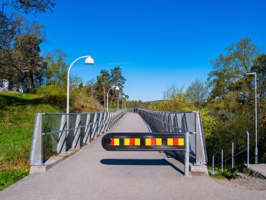 Çağdaş bir yaya köprüsünün üzerinde açık mavi gökyüzünün altında yeşil ve ağaçlarla çevrili bir bariyer var. Sahne bir sükunet ve doğa hissi veriyor..