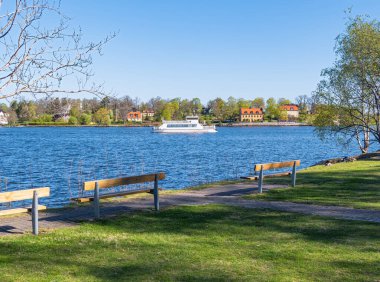 Stockholm, İsveç - 5 Mayıs 2025: Arka planda yemyeşil ve büyüleyici evlerle çevrili sularda yüzen bir teknenin yer aldığı sakin bir göl manzarası. Sahilde sıralanan banklar rahatlatıcı..