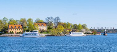 Stockholm, İsveç - 5 Mayıs 2025: Açık mavi gökyüzü altında tekneleri, yemyeşil alanları ve büyüleyici evleri olan manzaralı bir deniz manzarası.
