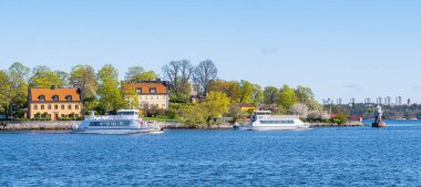 Stockholm, İsveç - 5 Mayıs 2025: Tekneleri, yemyeşil bahçeleri ve büyüleyici evleri olan deniz manzaralı bir manzara. Gökyüzü berrak ve mavidir, sakin bir atmosferi yansıtır..