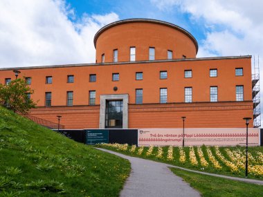 Stockholm, İsveç - 11 Mayıs 2025: Turuncu duvarları ve geniş pencereleri olan yuvarlak bir çatısı olan modern bir mimari bina. Ön planda çiçekli çimenli bir yamaç ve girişe giden bir patika yer alıyor. Bui 'de bir pankart sergileniyor