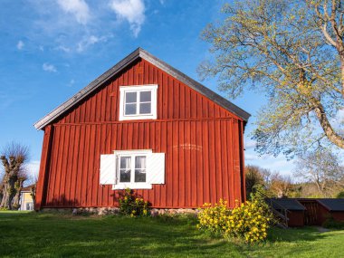 Stockholm, İsveç - 2 Mayıs 2025: açık mavi gökyüzüne karşı beyaz süslemeli büyüleyici kırmızı ahşap bir ev. Ev eğimli bir çatıya sahiptir ve yeşillik ve çiçek açan sarı çiçeklerle çevrilidir..