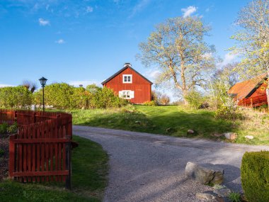 Bereketli yeşilliklerle çevrili kırmızı ahşap bir ev, eve giden çakıl yolu ve açık mavi bir gökyüzünün olduğu pitoresk kırsal bir manzara. Manzara sakin ve davetkar, bir ağaç ve bir lamba direği var..