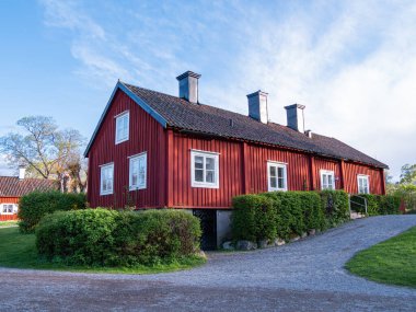 Stockholm, İsveç - 2 Mayıs 2025: Çatı eğimli, yemyeşil ve çakıl yollarıyla çevrili büyüleyici kırmızı ahşap bir ev. Ev, açık mavi gökyüzüne karşı dizilmiş birden fazla pencere ve baca yığınına sahip..
