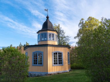 Stockholm, İsveç - 2 Mayıs 2025: Yeşilliklerle çevrili, tepesinde kubbeli bir çatısı ve bayrağı olan büyüleyici, ahşap bir bina. Yapıda geniş pencereler ve açık mavi gökyüzüne karşı kurulmuş sıcak sarı bir dış yapı yer alıyor..