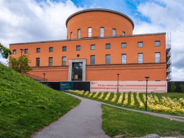 Stockholm, İsveç - 11 Mayıs 2025: Kırmızımsı-turuncu cepheli, yuvarlak kaplamalı modern bir mimari bina. Yapı yeşil çimlerle çevrili ve ona uzanan bir patika, önünde de bir çiçek yatağı var. İnşaat alanı.