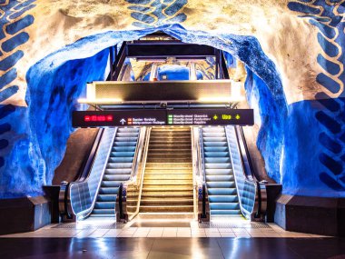 Stockholm, İsveç - 3 Haziran 2025: Sanatsal mavi kaya oluşumları ve aydınlatılmış yürüyen merdivenleri olan çarpıcı bir metro istasyonu manzarası. Modern tasarım, mağara benzeri duvarların doğal dokusuyla çelişiyor..