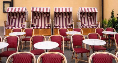 Sarı bir duvara dayanmış, ahşap çerçeveli çizgili plaj sandalyeleri. Önde, kırmızı hasır sandalyelerle çevrili yuvarlak mermer tepeli masalar var, rahat bir açık hava oturma alanı oluşturuyor..