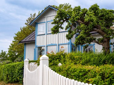 Vaxholm, İsveç - 4 Haziran 2025: Yeşillik ve beyaz çitlerle çevrili mavi aksanlı büyüleyici ahşap bir ev. Ev büyük pencerelere ve eğimli bir çatıya sahiptir..
