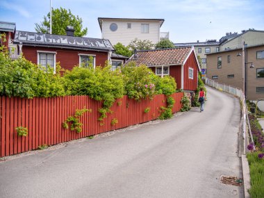 Vaxholm, İsveç - 4 Haziran 2025: Renkli evler ve yemyeşil alanlarla kaplı dar bir caddenin manzarası. Ön planda kırmızı ahşap bir çit ve büyüleyici kırmızı bir ev var..