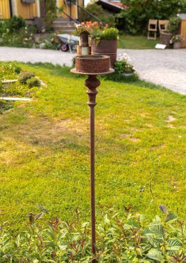 Yıpranmış metalden yapılmış, yeşil bir bahçede dimdik duran köy meşalesi standı. Meşale tutacağı boş, yemyeşil bitkilerle çevrili ve arka planda bir çakıl yolu var..