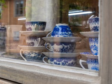 Bir mağaza vitrininde sergilenen dekoratif porselen çay fincanları ve çay tabakları koleksiyonu. Bardaklarda karmaşık mavi ve beyaz desenler var, klasik bir estetik sergiliyorlar..