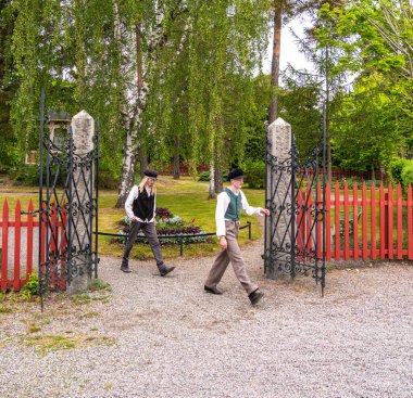 Sodertalje, İsveç - 6 Haziran 2025: Geleneksel İsveç kıyafeti giymiş iki genç yeşil çitlerle çevrili bir bahçe ortamında süslü bir kapıdan geçerek yürüyorlar.