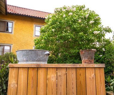 Vaxhom, İsveç - 4 Haziran 2025: Üzerinde iki kap bulunan taşralı ahşap bir çit: parlak metal bir küvet ve yıpranmış bir kova. Arkalarında, yemyeşil bir çalı ve beyaz çiçekler vardı..