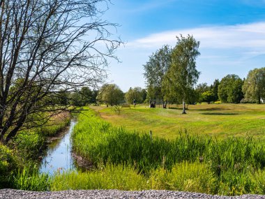 Stockholm, İsveç - 13 Haziran 2025: Ağaçları ve küçük bir deresi olan yemyeşil bir arazi. Gökyüzü açık ve mavi, huzurlu bir açık hava manzarası yaratıyor..