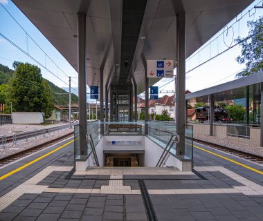 Yeraltına inen merdivenleri, cam ve metal yapılarla çevrili modern tren istasyonu platformu ve her iki tarafında da raylar var. Arka planda ağaçlar ve binalar görülebilir..