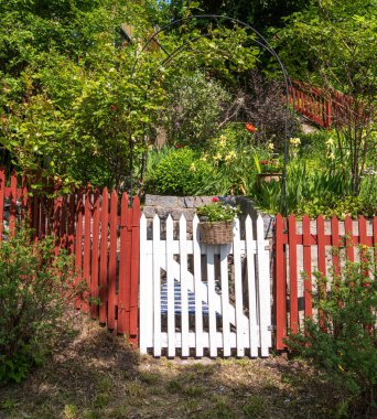 Beyaz çitli, kırmızı aksanlı, yemyeşil ve renkli çiçeklerle çevrili büyüleyici bir bahçe kapısı. Sahne huzur dolu ve davetkar, sükunet hissi uyandırıyor..