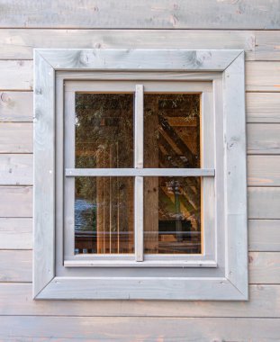 Ağaç desenli ahşap bir pencere çerçevesi, ağaçları yansıtan ve dışarıda ahşap bir yapı. Çerçeve açık gri renge boyanmış, ahşap duvarı tamamlıyor..