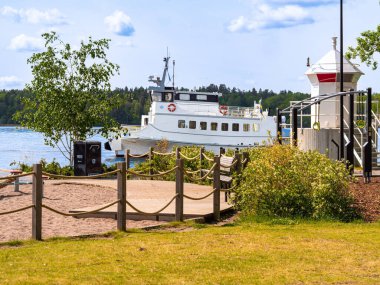 Stockholm, İsveç - 10 Haziran 2025: ahşap bir geçide kenetlenmiş beyaz feribotlu bir limanın manzarası. Bereketli bir yeşil alan, kumlu bir plaj ve açık mavi gökyüzünün altında bir deniz feneri ile çevrelenir..
