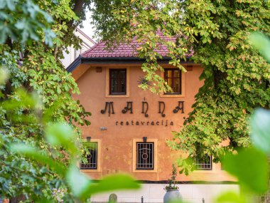 Ljubljana, Slovenya - 22 Temmuz 2025: Kırsal turuncu dış ve sanatsal harflere sahip yemyeşil ağaçlarla çevrili büyüleyici bir restoran cephesi. Sahne samimi ve davetkar bir atmosferi çağrıştırıyor, yemek için mükemmel..