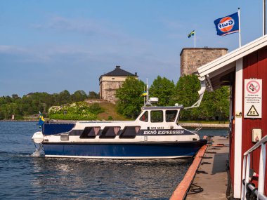 Vaxholm, İsveç - 4 Haziran 2025: Arka planda yemyeşil ve tarihi binalarla çevrili İsveç bayraklarıyla bir rıhtıma 'Ek-Island Expressen' adlı küçük bir yolcu feribotu yanaştı.