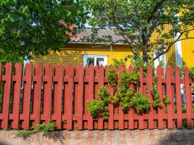 Vaxholm, Sweden - 4 June 2025: Red wooden fence with green foliage and yellow house in background.