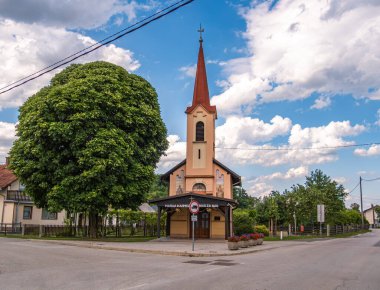 Odranci, Slovenya - 24 Temmuz 2025: Slovenya 'da İzakovci' de kırmızı bir çan kulesi ve önünde bulutlu mavi bir gökyüzünün altında bir ağaç bulunan küçük bir kilise.