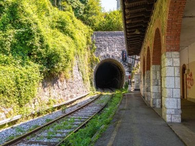 Yeşillik ve taş duvarlarla çevrili bir demiryolu tüneli girişi ve tünele giden izler. Sahnede kemerli mimarisi ve işaret ışığı olan bir platform yer alıyor..
