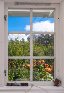 Renkli çiçekleri ve mavi gökyüzünün altındaki ağaçları gösteren bir pencereden bak..
