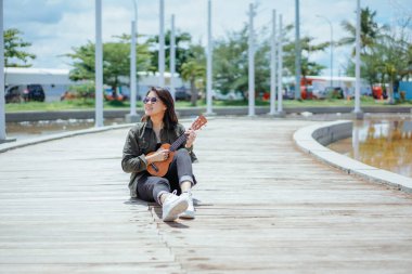 Ceket ve açık havada siyah kot giyen güzel bir Asyalı kadının Ukulele 'sini oynuyorum.