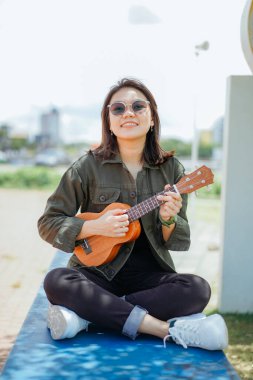 Ceket ve açık havada siyah kot giyen güzel bir Asyalı kadının Ukulele 'sini oynuyorum.