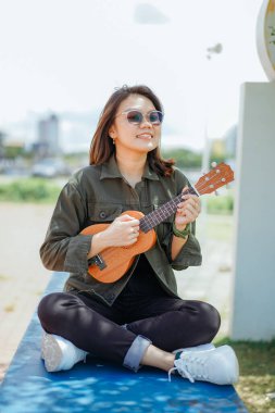Ceket ve açık havada siyah kot giyen güzel bir Asyalı kadının Ukulele 'sini oynuyorum.