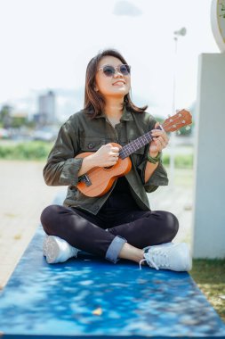 Ceket ve açık havada siyah kot giyen güzel bir Asyalı kadının Ukulele 'sini oynuyorum.