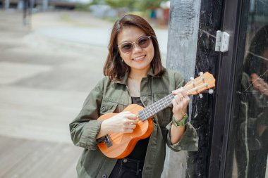 Ceket ve açık havada siyah kot giyen güzel bir Asyalı kadının Ukulele 'sini oynuyorum.